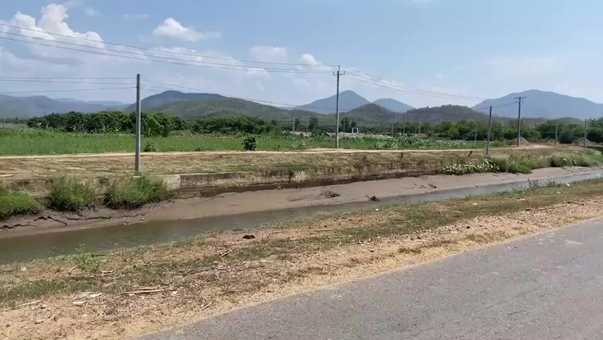 The camera focuses on a narrow, muddy canal beside a rural road. The banks of the canal are largely bare earth, with scattered patches of grass. In the distance, a cultivated field is visible, bordered by trees and utility poles. The horizon is marked by rolling hills and mountains under a blue sky with scattered clouds. The scene appears peaceful and somewhat arid.
