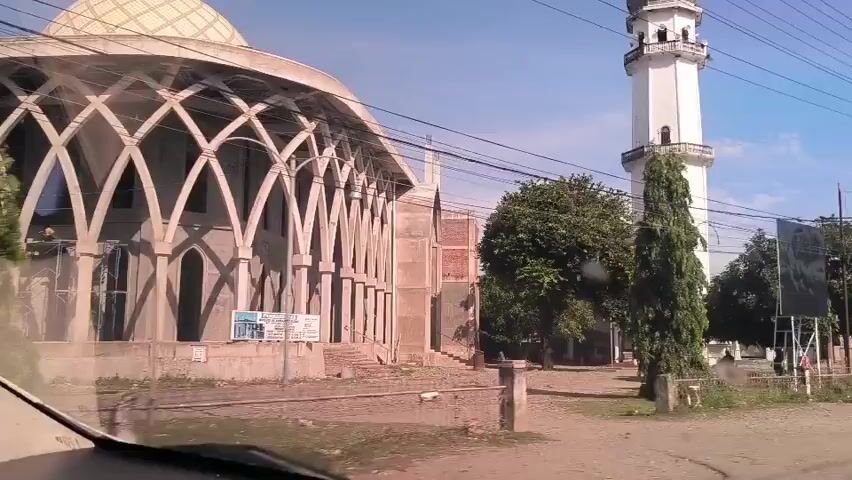 The video shows a car driving by a mosque under construction. The mosque has a dome and a tall minaret. The facade is composed of repeating arch shapes. There are power lines crisscrossing above the mosque, and a green street sign that reads "Banda Aceh" in one direction and "Jabal Ghafur" in the other. A person rides by on a motorcycle. The sky is bright blue.