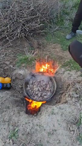 A pot filled with chunks of meat simmers over an open fire, set in a shallow earthen pit surrounded by dry brush and grass. The fire burns intensely, with orange flames licking around the bottom and sides of the pot. Steam rises from the meat inside the pot. Two individuals are partially visible, one's leg clad in Adidas clothing and the other's foot with a shoe holding a cooking utensil, watching the cooking process. A yellow toy truck sits on the ground nearby.