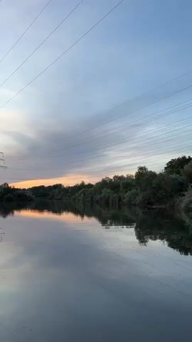 The video shows a serene natural landscape with a body of water in the foreground. The water reflects the sky and the surrounding trees, creating a mirror-like effect. Power lines stretch across the sky, and transmission towers are visible on the horizon. The sky is painted with the soft hues of sunset, with clouds transitioning from blue to shades of orange and pink. The overall impression is one of tranquility and natural beauty, juxtaposed with the presence of industrial infrastructure.