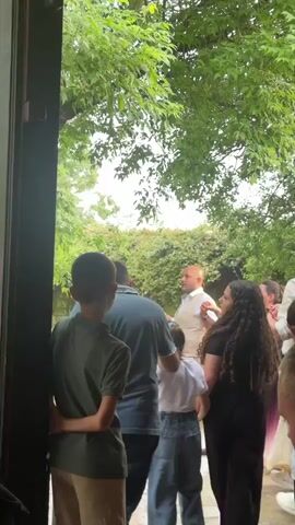 A low-angle shot captures a group of people dancing outdoors from a building's doorway. A young boy in a green shirt and khaki pants stands with his back to the camera, his hands clasped behind him. Behind him, a man in a blue shirt bends over slightly, interacting with a young girl in a white shirt and blue jeans. To the right of the man, a woman in a purple dress dances with a man in a white suit. Other individuals are partially visible, dancing in the background. The scene is set under a canopy of green trees, creating a festive atmosphere.
