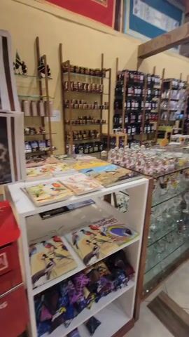 The camera pans slowly to the right to reveal the interior of a souvenir shop. Several shelves display neatly arranged rows of small jars, black bags, and other goods. In the center of the frame, a glass-topped counter showcases a variety of glasses, mugs, and other items. A salesperson in a black shirt stands behind the counter, facing away from the camera. A person in a patterned dress walks down the aisle in the distance. The floor has a light-colored tile with a large "X" pattern. Wooden beams support the ceiling, and the walls are painted in warm tones.