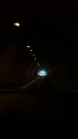 The video shows a view from inside a vehicle traveling through a dark tunnel. The tunnel has a curved ceiling with a row of lights illuminating the path. The road ahead is visible, with double white lines marking the lanes. At the end of the tunnel, there is a bright light, suggesting the exit. The interior of the vehicle, including the dashboard, is partially visible in the foreground. The overall ambiance is dark and enclosed, with the light at the end of the tunnel offering a sense of progression.