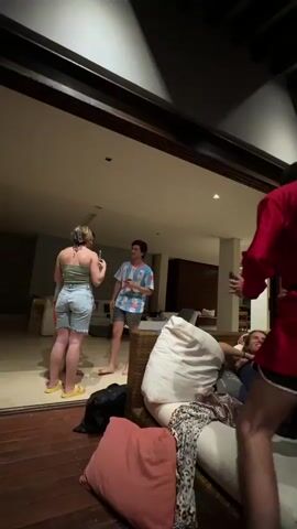 The video shows a group of young adults at a casual gathering in what appears to be a vacation home. Indoors, three people are gathered near a large mirror, while a person in a red robe walks toward a cushioned seating area where two people are lounging. The scene then shifts to an outdoor space with exposed wooden beams and what appears to be a deck. A table with a red plastic cup sits between two large speakers. Two men are standing at the table, one wearing red shorts and a white t-shirt with "PUTO" written on it. A person lies on the couch, eating pizza from a blue plate. The setting suggests a relaxed, social atmosphere.