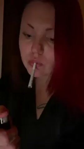 Here's a concise description of the video:

A woman with bright red hair lights a cigarette with a lighter and takes a drag. She’s wearing a dark robe or shirt, and has tattoos on her hands and neck. The setting is dimly lit, possibly indoors.