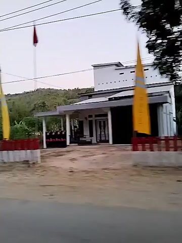 The video shows a view from a moving vehicle. On the right side of the road, there is a partially constructed white building with open-air levels supported by columns. A flag with green, yellow, and red stripes is attached to a pole in front of the building. Lush green vegetation and trees are visible around the building and in the background, with a gently sloping hill covered in greenery. Power lines are strung across the sky. In the foreground, there is a paved road. The lighting suggests it is either early morning or late afternoon.