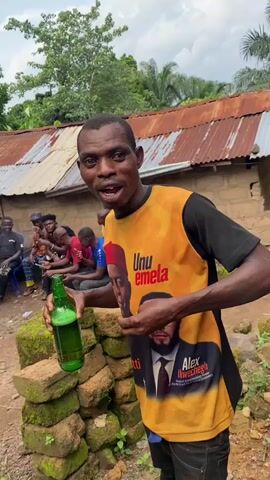 A man in a yellow t-shirt with pictures of two men on it holds a green bottle of liquid in his left hand and gestures with his right hand. He is standing outdoors in front of a building with a rusted corrugated metal roof. Several other men are seated in the background. The ground is dirt and there are some green plants and moss-covered bricks. The sky is overcast.