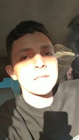 A young man takes a selfie in the front passenger seat of a car. A older man with a beard and a dark green beanie leans into frame next to him. The camera then shows the driver and the road ahead through the windshield, bathed in sunlight. The interior of the car and the buildings and scenery outside are visible. The car is driving on a road, and buildings are visible in the background. The driver is steering. The car radio is on.