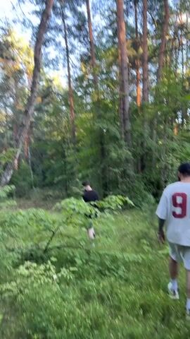 Two individuals walk through a dense forest. The person in the foreground walks away from the camera, wearing a white t-shirt with the number 91 on the back, white shorts, and white sneakers. This person has tattoos visible on their left arm and is wearing a baseball cap. The second individual, further ahead and slightly to the left, walks away from the camera wearing a dark t-shirt and shorts. The environment is characterized by tall trees, lush green foliage, and overgrown grass, suggesting a natural, wooded area during daylight hours. The sunlight filters through the trees, illuminating parts of the scene.