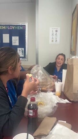 Three women are sitting at a wooden table, eating from plastic bags. The bags appear to contain seafood, possibly shrimp. There are also drinks, including what appears to be soda and juice. One woman is holding a piece of food, possibly a potato or corn. They seem to be engaged in conversation. The setting appears to be a break room or office, with a bulletin board on the wall that says "Employment".