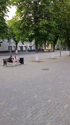 A video shows a street scene with people walking and sitting. In the foreground, a paved walkway is visible. A bench is situated on the walkway, occupied by two individuals, one appearing to be a young woman and the other a young man. Further down the walkway, another bench is occupied by a young man. Across the street, a group of young people are seen walking. Trees line the street, and buildings are visible in the background. The overall impression is of a public space during daylight.