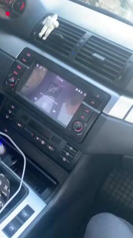 The video provides a first-person perspective from inside a car. The camera pans across the dashboard. Visible on the dashboard are a car stereo with a screen displaying an image, air vents, and a small white astronaut figurine. Further along the dashboard, a pink sticker with text that appears to be in Cyrillic script and a crown symbol is present. A plush pink octopus toy is also visible on the dashboard. Outside the car, through the windshield, a street scene with buildings and a few people can be observed. The lighting suggests it is daytime, possibly late afternoon or early evening. The video has an overlaid timestamp at the beginning.