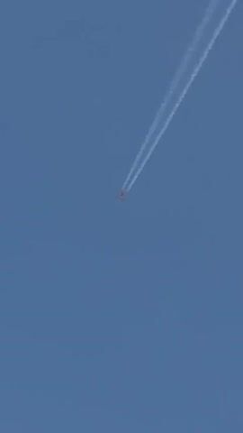 The video shows an airplane flying across a clear blue sky, leaving a contrail.  The plane is small and distant. The scene then shifts to a view from under an overhanging roof.  The sky is now a lighter blue, with some pink-tinged clouds and the moon visible. Below the sky is a view of rooftops and trees. Mountains are visible in the distance.