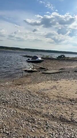 The video shows a view of a riverbank with several boats. In the foreground, there is a sandy and pebbly beach. The water is choppy and a boat is partially submerged on the left side. A wooden ramp leads to a yellow and white boat docked on the right side of the beach. In the distance, across the river, a treeline is visible under a blue sky with scattered clouds. Another boat, possibly a barge, is seen further in the distance on the river. There are no visible people in the video.