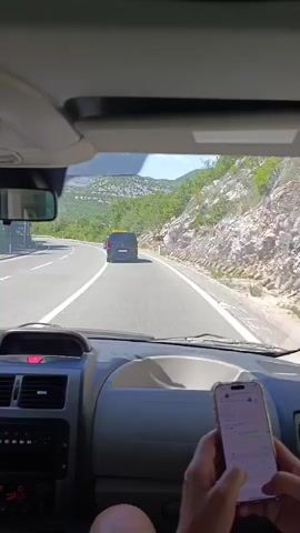 The video is filmed from inside a vehicle, looking forward through the windshield. A person's hands are visible in the foreground, holding a smartphone. The smartphone screen displays a messaging application, with text and icons visible. The view through the windshield shows a paved road with white lane markings, curving to the left. A black van is driving ahead in the same lane. On the right side of the road, there is a rocky hillside with sparse green vegetation. A yellow road sign with black text and symbols is visible on the right side of the road. In the distance, rolling hills covered in green trees and some rocky outcrops are visible under a bright, sunny sky.