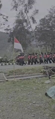 A group of people in uniform are marching in formation on a road. They are wearing red vests over dark uniforms and some are carrying what appear to be rifles. They are marching past a flag pole with a red and white flag flying on it. The environment appears to be outdoors in a rural or mountainous area with trees and hills in the background. There are also motorcycles parked on the side of the road.