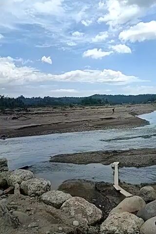 The video shows a river flowing through a dry, rocky, and sandy landscape. The river has several shallow channels with exposed sandbars. The sky is partly cloudy with white clouds against a blue sky. In the distance, there are hills covered with trees. There are a few people walking in the distance. The foreground shows large rocks and sand. There is a makeshift wooden structure in the river. The overall impression is of a natural landscape, possibly a riverbed during a dry season.