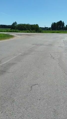 The video begins with a first-person perspective of a cyclist on a paved road, showing the handlebars with a bright yellow grip. The road surface is cracked and appears to have some patches. The surrounding environment is rural, with green fields and trees visible under a clear blue sky. As the video progresses, signs appear by the side of the road, one of which is yellow with text in a foreign language and an arrow pointing to the left, and another blue sign with text and an arrow. The cyclist continues to ride, and the camera briefly focuses on the face of a young man wearing a baseball cap and a backpack, looking directly at the camera. The video concludes with a shot of trees and a clear sky.