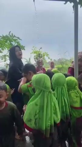 Children, caked in mud, gather near a water spout. Some are in green hijabs and shirts, while others wear red-trimmed green shirts. They stand in a puddle of water, some looking down, others at the water spout. Adults surround them, including women in hijabs and a man in a black cap. One woman points towards the water spout, possibly instructing the children to wash off the mud. The scene unfolds outdoors, with foliage and a cloudy sky visible in the background.