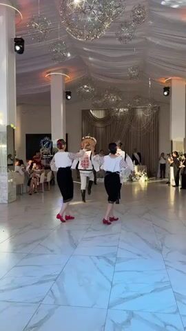 A group of dancers in traditional attire perform a dance in what appears to be a banquet hall or reception venue. Four dancers are visible in the foreground, facing away from the camera. They are all wearing white blouses with embroidered sleeves, black skirts, and red shoes. Two of the dancers are wearing their hair up with colorful floral accents. The dancers in the middle are wearing white pants and vests with intricate embroidery, and tall black boots. One of these dancers is holding a decorated bread over their head. The floor is a polished marble with a geometric pattern. In the background, there are tables with floral arrangements, draped curtains, and hanging decorative lights. Other guests are visible in the background, some seated at tables and others standing.
