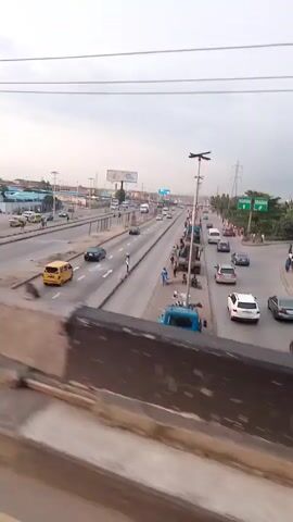 The video shows a view from an elevated position looking down at a busy highway. Numerous cars, buses, and taxis are traveling in both directions. There are people walking on the side of the road and standing near market stalls. Overhead power lines and streetlights are visible. The sky is overcast, and the overall lighting suggests it is daytime.
