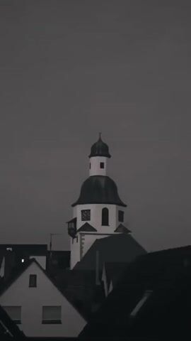A black and white video shows a church tower with a black dome and a small spire against a dark sky. There are also rooftops of houses in the foreground and middle ground. The clock on the tower is visible.