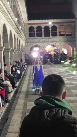 A fashion model walks down a tile runway in an outdoor courtyard. The model wears a blue and silver dress. A crowd of people sits on one side of the runway. On the other side is a man with a green hooded jacket. More people stand with cameras and lights at the end of the runway. The environment has stone arches and gardens. A drone flies above the model, filming.