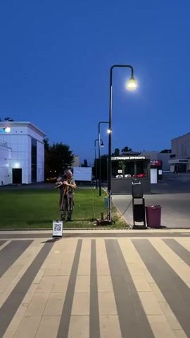 A man dressed in a costume stands in a grassy area next to a sidewalk. He holds a long, thin object that resembles a staff or pole. The man appears to be performing or posing. There is a lamppost nearby that is illuminated. Behind the man is a building with white walls and a large blue sign. Further in the background, there are other buildings and a small kiosk. The scene is lit by artificial lights, suggesting it is either dusk or nighttime. The ground in the foreground is paved with dark and light striped tiles.