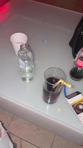 The video shows a table with drinks and a cigarette pack, then pans up to show a nightclub scene. The table holds a glass of cola with a yellow striped straw, a small open bottle of water, and a small white paper cup that looks to contain a white solid substance. The cigarette pack is partially visible at the edge of the table. As the camera lifts and pans around, it reveals a nightclub interior. People are seen sitting at tables and standing, some wearing casual clothing like t-shirts with printed designs. A large video screen is visible on the back wall, displaying a stylized logo. The lighting in the club is colorful, mainly purple and blue, and is consistent with a nightlife setting. The furnishings include cushioned chairs and tables that appear to glow with colored light. The ceiling is decorated with a geometric pattern. There is music playing in the background.