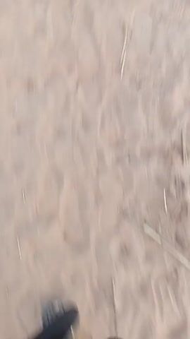 The video shows two men walking on a sandy surface, away from the camera. One man is wearing a burnt orange robe, while the other is wearing a white robe. The ground is tan-colored and appears to be sandy with scattered twigs and straw. Some trees are visible in the background. The camera is low to the ground and follows the men as they move. The video seems to be shot by someone walking behind them.