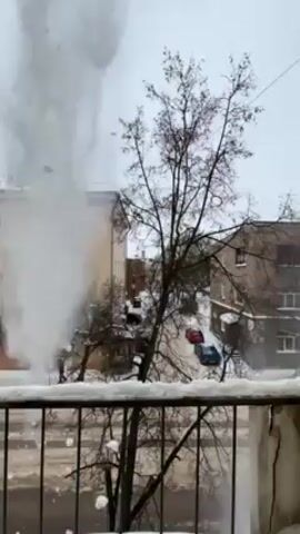 The video shows a burst pipe spewing water, debris, and steam into the air. The view is from a balcony overlooking a street covered in snow. There are buildings on either side of the street, and a few parked cars are visible. A person is walking on the sidewalk near the water plume.  The sky is overcast, and the general scene suggests a cold, winter day.