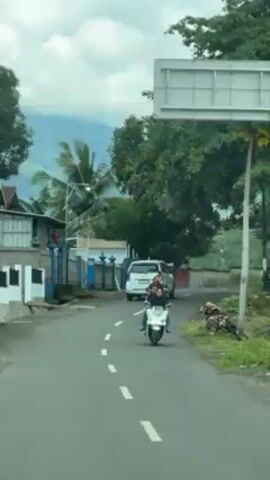 The video shows a view from inside a vehicle, likely a car, driving on a paved road through a rural area. In the foreground, the road is marked with dashed white lines. A white minivan drives ahead. Several people ride scooters on the road. Buildings and greenery are visible on either side of the road. Mountains can be seen in the distance. The sky is overcast.