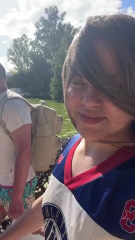 Here's a detailed description of the video:

The video appears to be shot outdoors near a lake or pond. The water is green, possibly due to algae. The weather looks sunny and bright.
The video starts with a close-up of a person smiling at the camera. They are wearing a blue and red jersey.
The camera then pans to show a man walking along a paved path next to the water. He is wearing a white t-shirt, a tan backpack, and patterned shorts that have an ice cream cone design on them. He is carrying a white plastic bag in his hand.
The man walks along the path, with the camera following him. The path is made of red bricks. There are trees and shrubs visible in the background. The sun is shining brightly.