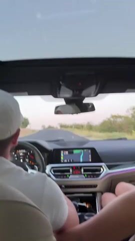 The video shows the interior of a car from the perspective of a passenger sitting behind the driver. The driver, a person wearing a white cap, is visible from the back of their head and shoulders. Their left hand is on the steering wheel, which shows a digital speedometer reading around 112. The car has a modern dashboard with a large infotainment screen displaying a navigation map. The passenger in the front seat, on the right, has their arm resting on the center console, and their hand is visible. The dashboard has ambient lighting. The car appears to be driving on a paved road, with trees and vegetation visible on either side. The sky is light and appears to be either dusk or dawn. The overall scene suggests a couple or friends traveling by car on a highway.