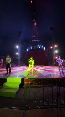 A low-angle shot captures a circus stage, bathed in bright lights, under a big top tent. A young person in a sleeveless yellow-green leotard, stands atop a unicycle, holding a microphone and speaking. A man in a white shirt with elaborate embroidery, and dark pants stands to the left, while a woman in a white, sparkly costume with high boots stands with her hand on her hip to the right. A row of empty seats is visible beyond the stage to the left. The stage is decorated with a red and white starburst pattern, and lit with alternating white, blue, and red lighting. A green-lit set of steps lead up to the stage on the left. The stage also has metal railing in the foreground. A large structure with spotlights and lights is centered and suspended from the tent.
