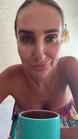A close-up, POV shot shows a woman with dark hair pulled back, her face angled down towards a teal mug of dark liquid. She is wearing a strapless top with a floral pattern. Her eyes are open and directed at the mug as she brings it to her lips to drink. The background is a textured wall with a decorative gold light fixture on the right side.