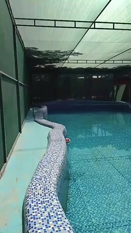 A video shows a swimming pool. The pool is covered by a white canopy supported by a metal frame. The walls of the pool area are dark green and the pool itself is tiled with small blue and white squares. There are lounge chairs and tables visible under a covered area in the background.