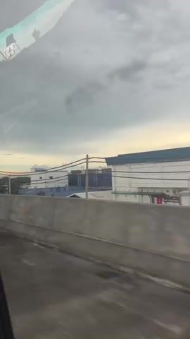 The video shows a view from inside a vehicle looking out at an industrial area with buildings and overcast skies. A concrete barrier runs along the side of a road in the foreground. In the distance, there are white and blue industrial buildings with power lines running across them. The sky is cloudy with a hint of sunlight breaking through. There are no people visible in the video.