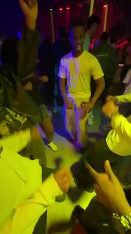 People are dancing and playing music in a nightclub. There are several people dancing on the floor, including a man in a white t-shirt and light pants. A woman in a black top and denim skirt is also dancing. A man is playing bongos, wearing a pink furry hat. The lighting in the club is colorful and dynamic.