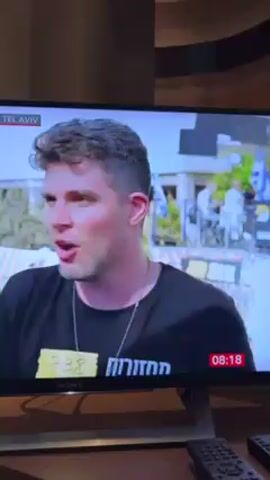 A man in a black T-shirt with writing on it is being interviewed on television. He is standing outside and speaks in earnest. The television is a Sony brand, and there is a remote control on the surface below it. A timecode in the corner of the screen indicates 08:18. The location is identified as Kings Square, Tel Aviv.