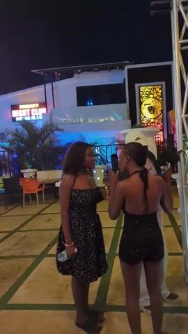 Two young women are standing in an outdoor paved area at night. The woman on the left wears a black strapless dress with white polka dots and holds a water bottle. The woman on the right wears a black halterneck top and black shorts. They are facing each other and appear to be talking. In the background, there is a building with a sign that reads "NIGHT CLUB" illuminated in red and white. There are tables and chairs outside the club, along with palm trees and decorative lighting. A metal structure is visible on the right side of the frame. Another person, partially visible, stands between the two women, looking towards the woman on the right.