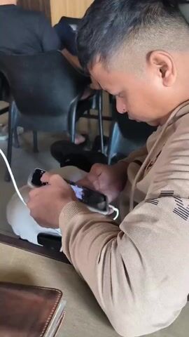 A man with short hair, wearing a tan hoodie, sits and uses two phones. One phone is held in his left hand, and the other is in his right. A white cable is plugged into the phone in his right hand, which is resting on a white object, possibly a phone case. The man's head is tilted down as he focuses on the screens. In the background, other people are seated, and chairs are visible. The environment appears to be indoors, possibly a cafe or waiting area, with a table in the foreground.
