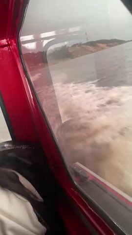 A first-person perspective video captures the view from inside a red boat cabin as it moves across the water. The camera is angled slightly downwards and to the right, looking out through the boat's window. Outside the window, foamy white water churns and splashes, indicating the boat's speed. Beyond the water, a hazy shoreline is visible with trees and a gently sloping hill dotted with what appear to be structures or cleared areas. The sky is overcast. Inside the cabin, part of a person's shoulder and dark hair are visible on the left side of the frame, suggesting they are seated and looking out the window. The interior of the cabin appears to be red, with some black and gray elements visible in the window frame and trim. The overall impression is a dynamic scene of travel on a body of water.