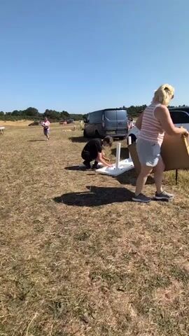 A young male wearing a black t-shirt and dark shorts is crouching in a grassy field, assembling a white plastic table. The table is upside down, and he is attaching or adjusting one of its legs. In the background, several cars and a van are parked. A white car with its trunk open is positioned behind the table. There are people in the background, some of whom appear to be standing near the cars. The sky is clear and blue, indicating a sunny day. The ground is covered in dry grass and some patches of bare earth. A cardboard box is visible on the trunk of the white car.