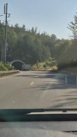 The video shows a first-person perspective of driving a vehicle on a highway towards a tunnel. The highway is paved with lane markings. On the left side of the road, there is a guardrail and a tall metal pole with equipment attached to it. The right side of the road also has a guardrail. The tunnel entrance is visible in the distance, framed by a dense forest covering a hillside. As the vehicle approaches the tunnel, two green trucks are seen on the sides of the tunnel entrance, spraying water over the opening, creating a visible mist. A white truck is in the process of entering the tunnel. The sky is clear and blue, and the lighting suggests it is daytime.