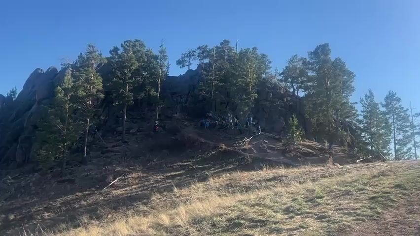 The camera is pointed up a hillside on a clear day, toward a rocky outcrop at the top. The slope is covered in dry grass, with a few rocks scattered on the hillside. The rocky outcrop at the top of the hill is populated with evergreen trees. There are several people on the outcrop, appearing as small and somewhat blurry figures. Blue material is draped in various places. In the distance a landscape is visible below and beyond the hilltop.