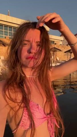 The video features a young woman in a pink polka-dot bikini posing in a body of water. She has long, wavy brown hair and is wearing a white beaded necklace. In the background, other people are visible swimming and relaxing in the water. The sun is setting, casting a warm glow over the scene and creating a peaceful atmosphere. The location appears to be a lake or beach, with buildings and vegetation visible on the shore in the distance. The woman's expressions range from a playful pout to a more relaxed smile, suggesting she is enjoying herself. The video captures a moment of leisure and relaxation during a beautiful sunset.