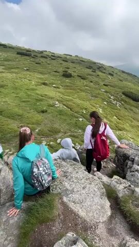 This media file captures a group of people hiking on a mountainous terrain in Cherna Mesta, Bulgaria. The scene is characterized by steep, grassy slopes dotted with rocks and low-lying vegetation. The sky is partly cloudy, with dramatic white clouds against a blue backdrop, suggesting it's daytime, likely late morning or early afternoon.

The subjects include several individuals, some actively hiking and others resting. In the foreground, two young women are visible. One, in a teal hoodie and a backpack with a floral pattern, is leaning on rocks, her hands on the stones. Beside her, another young woman in a white long-sleeved shirt and black leggings, wearing a bright red backpack, is looking down the slope. A third person, wearing a gray hoodie, is positioned further down the slope, appearing to be in the process of climbing or descending. In the background, a person in a white and green outfit is also visible on the trail.

The activity depicted is clearly hiking or trekking. The individuals are equipped with backpacks, suggesting they are on a journey through the mountains. The terrain is rugged, with a visible trail winding up the hillside. The overall mood seems to be one of outdoor adventure and possibly enjoyment of the scenic views, although the expressions are not clearly discernible.

Notable details include the rocky outcrops and the lush green of the mountainside, interspersed with patches of darker green shrubbery. The distant landscape reveals rolling hills and valleys covered in a dense forest, indicating a vast, natural environment. The weather appears pleasant for hiking, with a mix of sun and clouds. No visible text is present in the media.