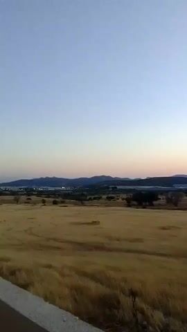 The video shows a panoramic view of a rural landscape at dusk. The sky transitions from a pale blue at the top to a soft orange and pink near the horizon. In the foreground, a field of dry, golden grass stretches across the scene. The grass appears somewhat windswept. In the middle ground, there are scattered trees and some structures that are not clearly identifiable. In the background, a range of dark mountains forms the horizon. The overall atmosphere is calm and serene, typical of a sunset in an open natural setting. There are no people or animals visible in the video. The lighting suggests that the sun has recently set or is about to set.