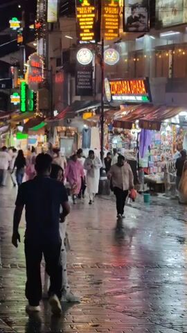 The video shows a bustling street scene at night, likely a market or shopping area. Numerous people are walking along a wet, paved pathway lined with shops and restaurants. Neon signs in various colors illuminate the buildings, advertising businesses like "Zimindara Dhabas," "Tree Spa," and "Punjabi Chinese South Indian." Some shops appear to be selling clothing or goods. A few individuals are in the foreground, some walking away from the camera, while others are engaged in conversation. The overall atmosphere is lively and commercial, with the wet ground reflecting the bright lights of the signs.